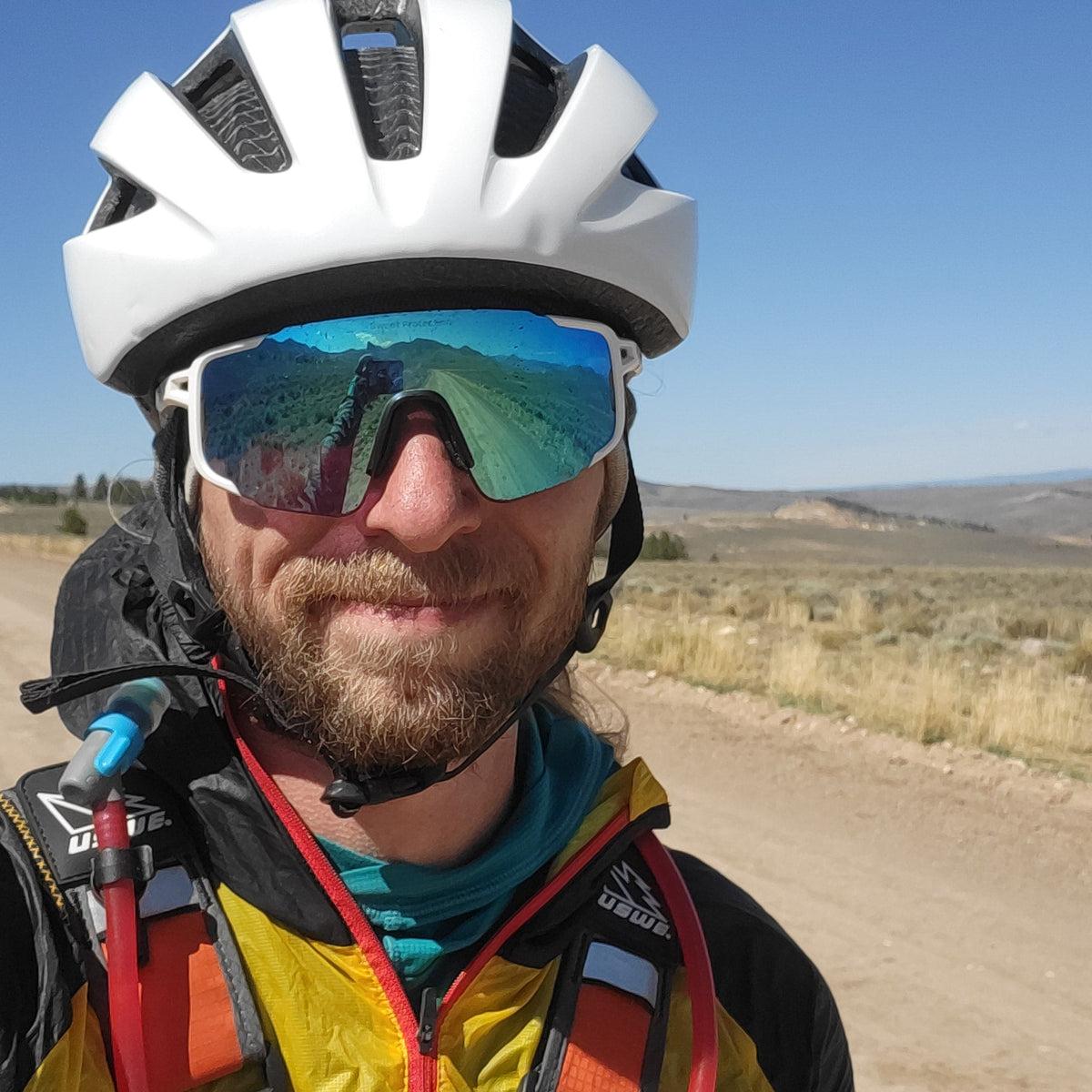 A close-up selfie of a cyclist outdoors on a dirt road, wearing a white helmet, mirrored sunglasses, a black and yellow jacket, and a hydration pack, with a desert landscape and blue sky in the background.