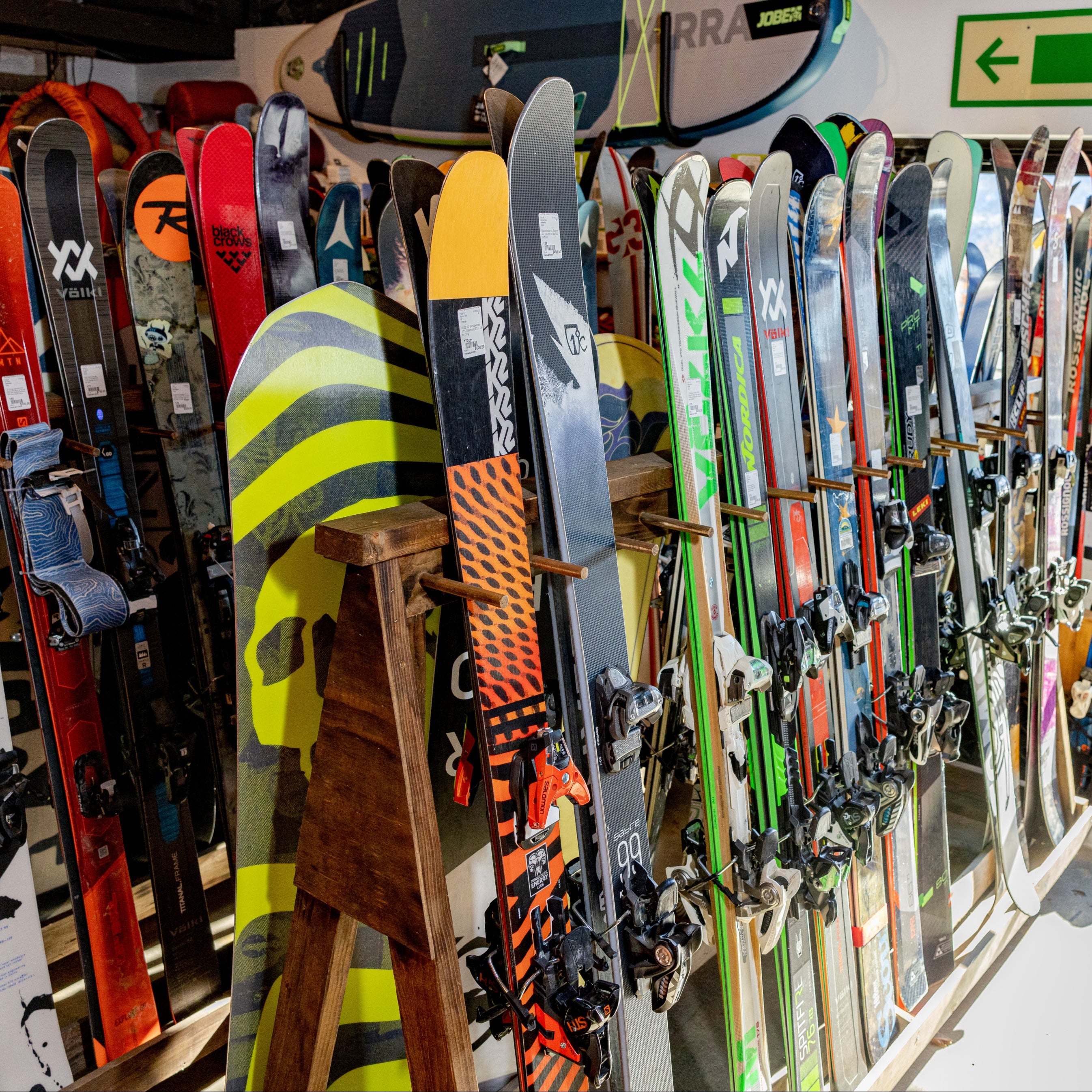 A retail display of various skis and snowboards hanging on racks inside a shop, with a green 'Mercy' directional sign above and a bright window in the background.