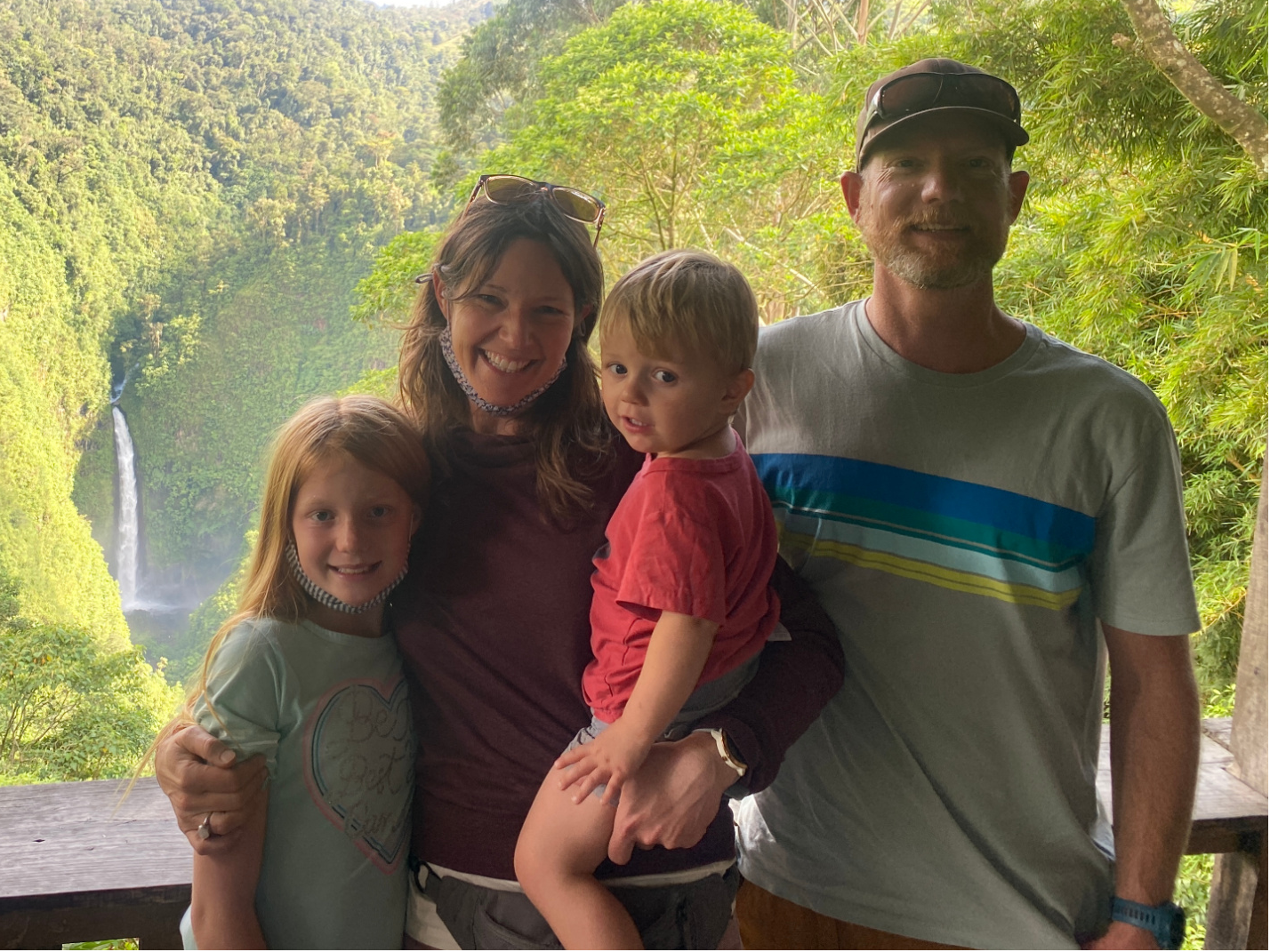 A family of four posing together on a wooden deck overlooking a lush green forest and a waterfall in the distance. The mother is wearing a burgundy top, the father wears a gray t-shirt with blue and yellow stripes, and the two children are in casual light-colored clothing.